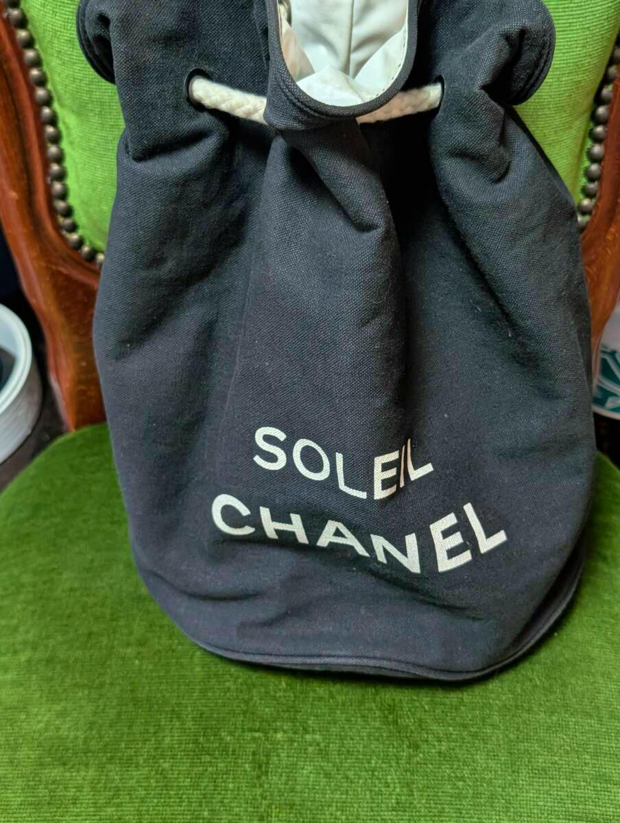 Front view of a black canvas CHANEL Soleil novelty backpack with 'SOLEIL' and 'CHANEL' printed on the front, placed on a green chair.