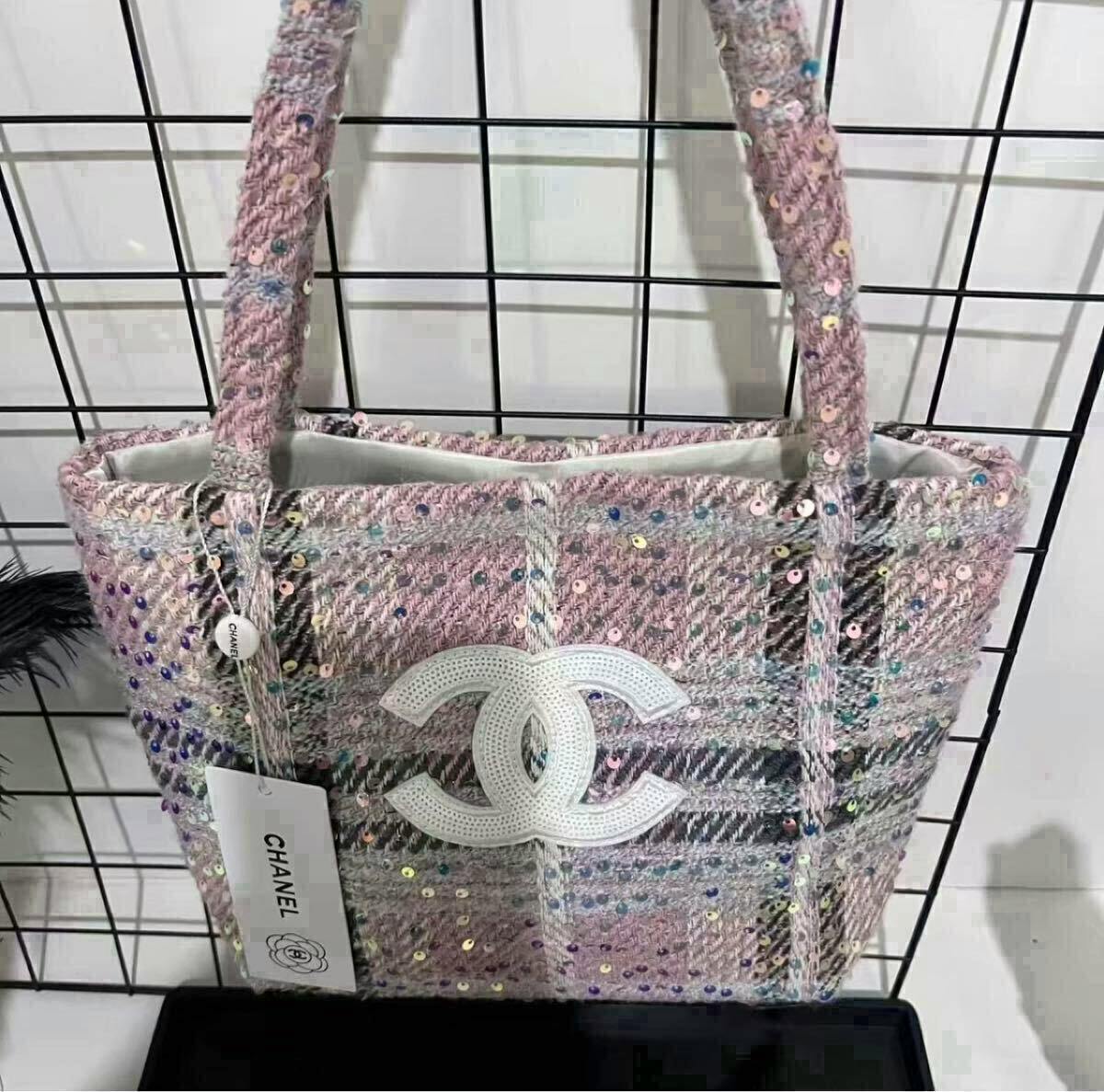 A pale pink tweed check-pattern Chanel tote bag with sequins and a white interlocking CC logo on the front, shown hanging by its handles.