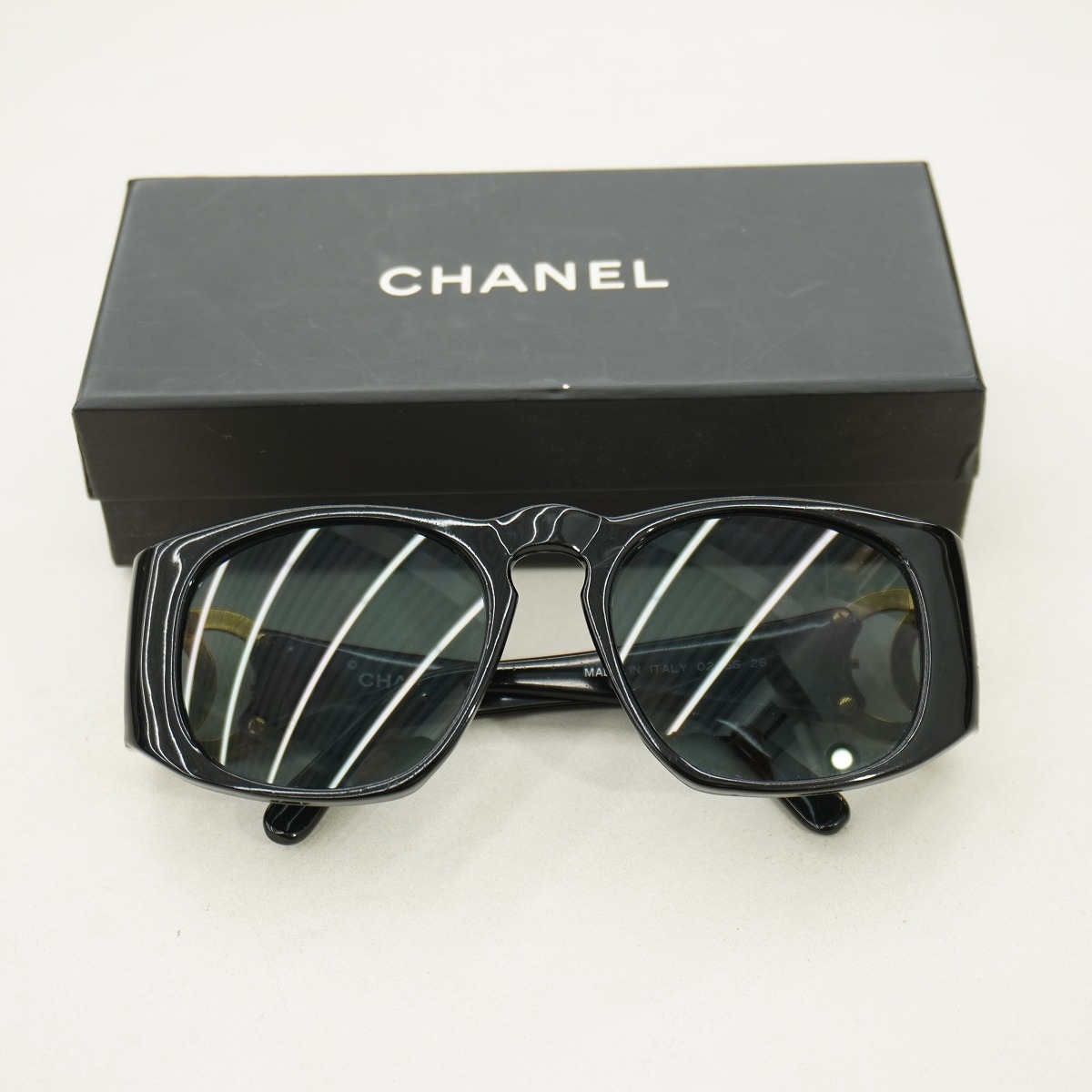 Black CHANEL sunglasses with dark lenses displayed in front of a CHANEL box on a white surface.