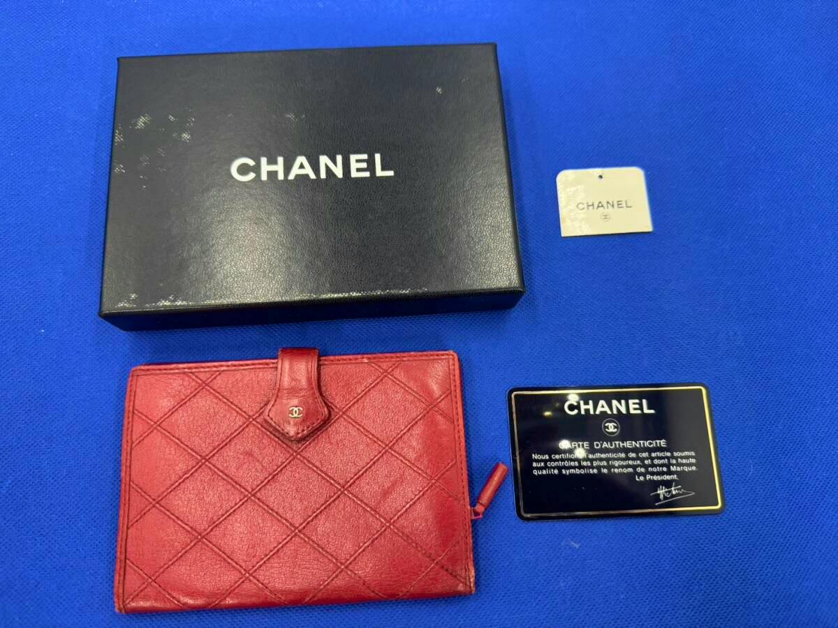 Red CHANEL bi-fold lambskin wallet with gold CC clasp, shown with its black CHANEL box and authenticity card on a blue background.