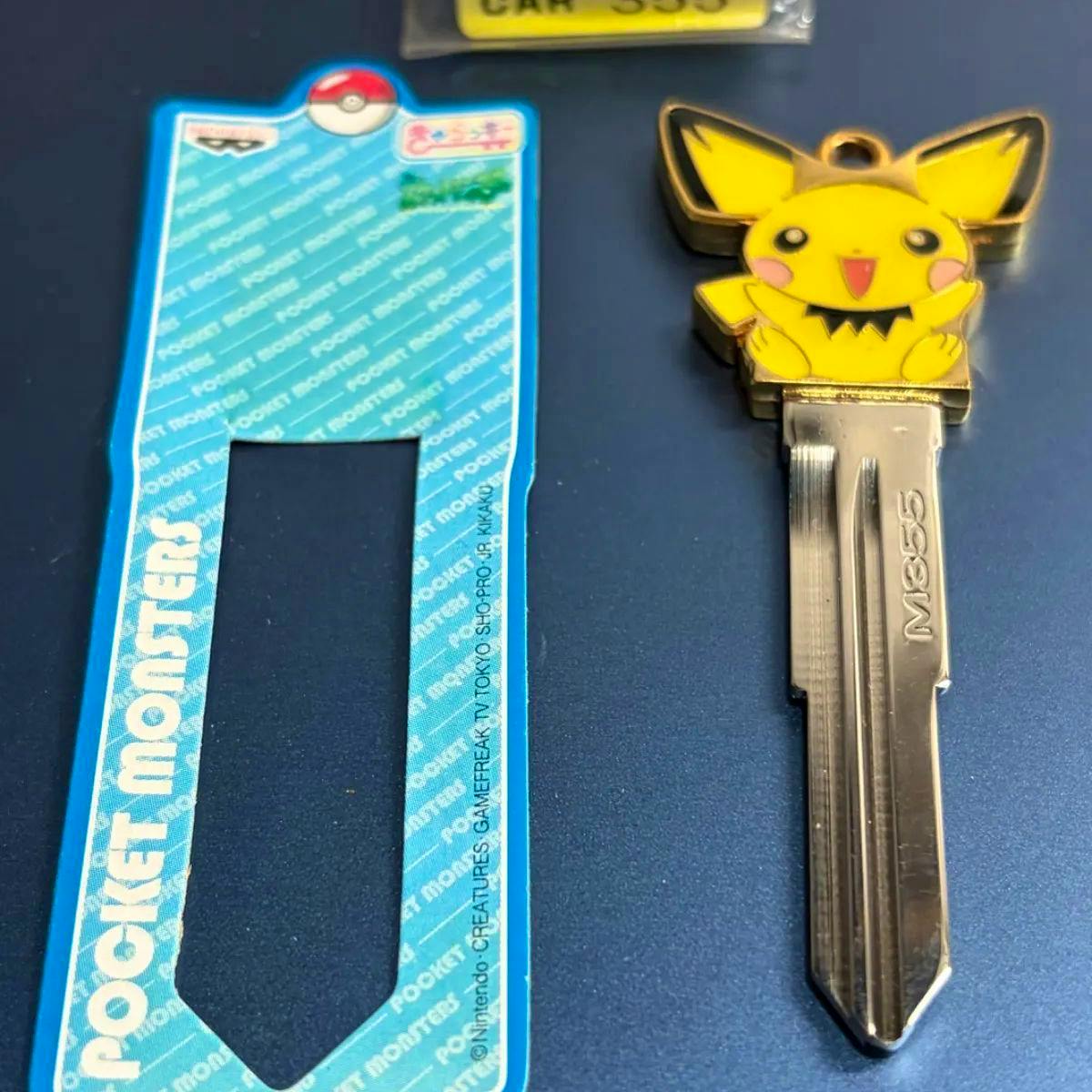Metal car key cover with a yellow Pikachu head attached to a key blank stamped "M355", shown beside a blue Pocket Monsters backing card.