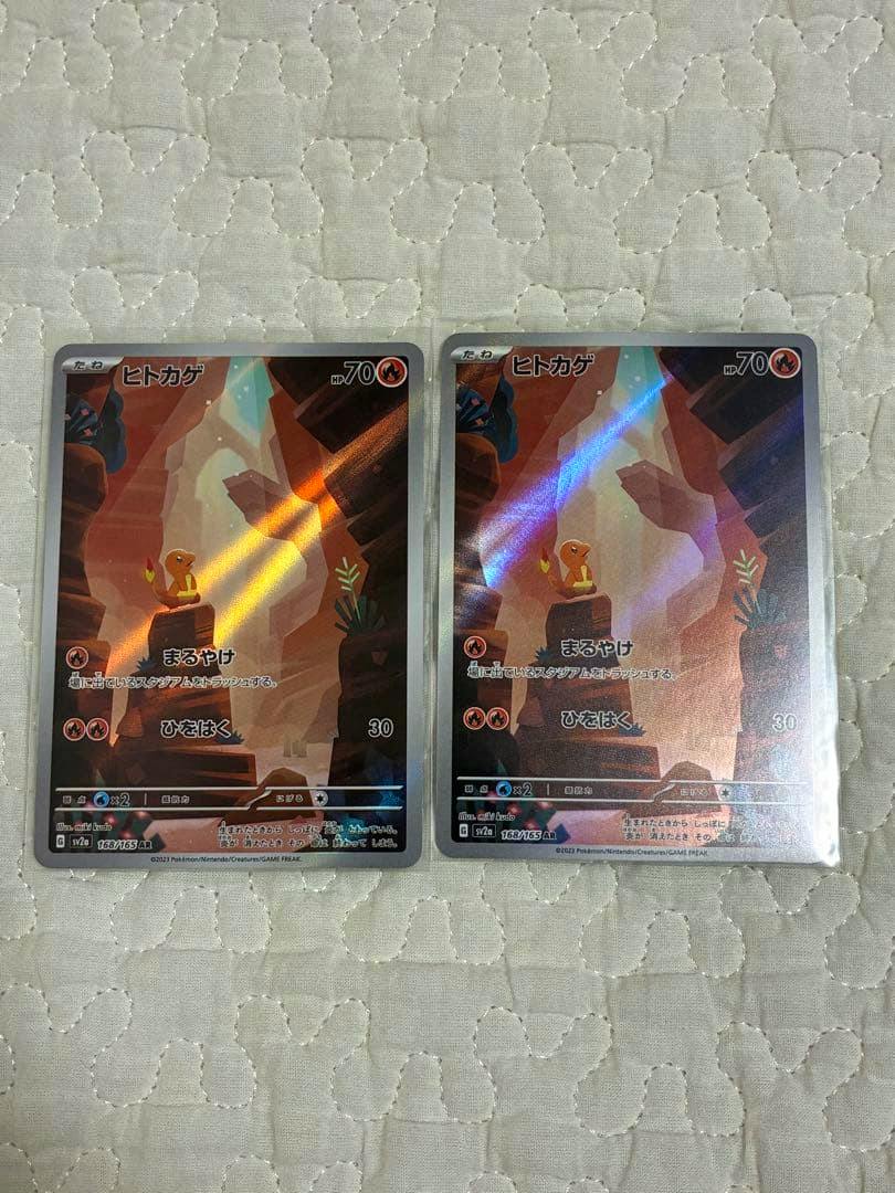 Front sides of two ヒトカゲ (Charmander) Pokémon trading cards with holographic foil shown side by side on a quilted fabric.