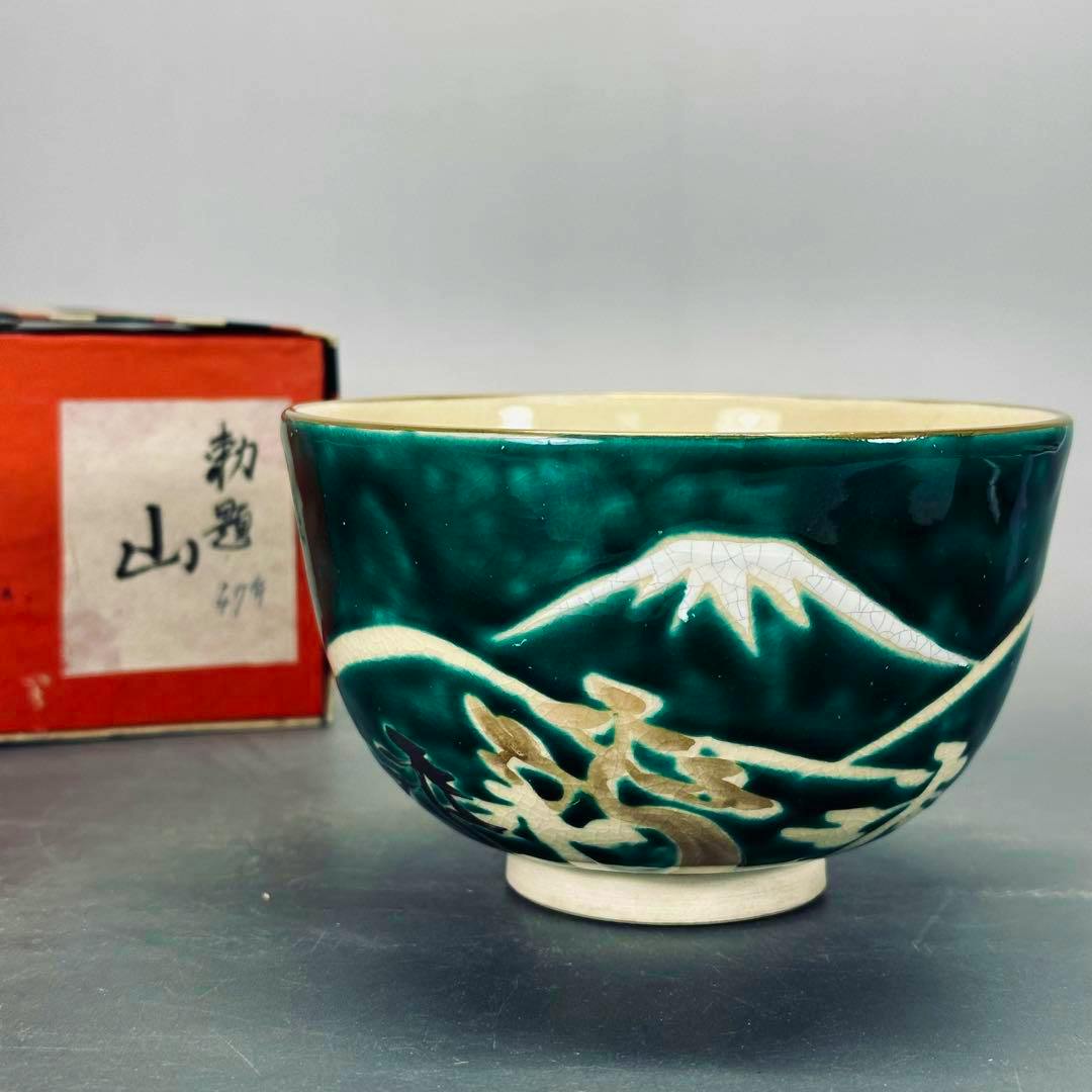 A green-glazed matcha bowl with a white mountain motif and its red box partially visible behind it.