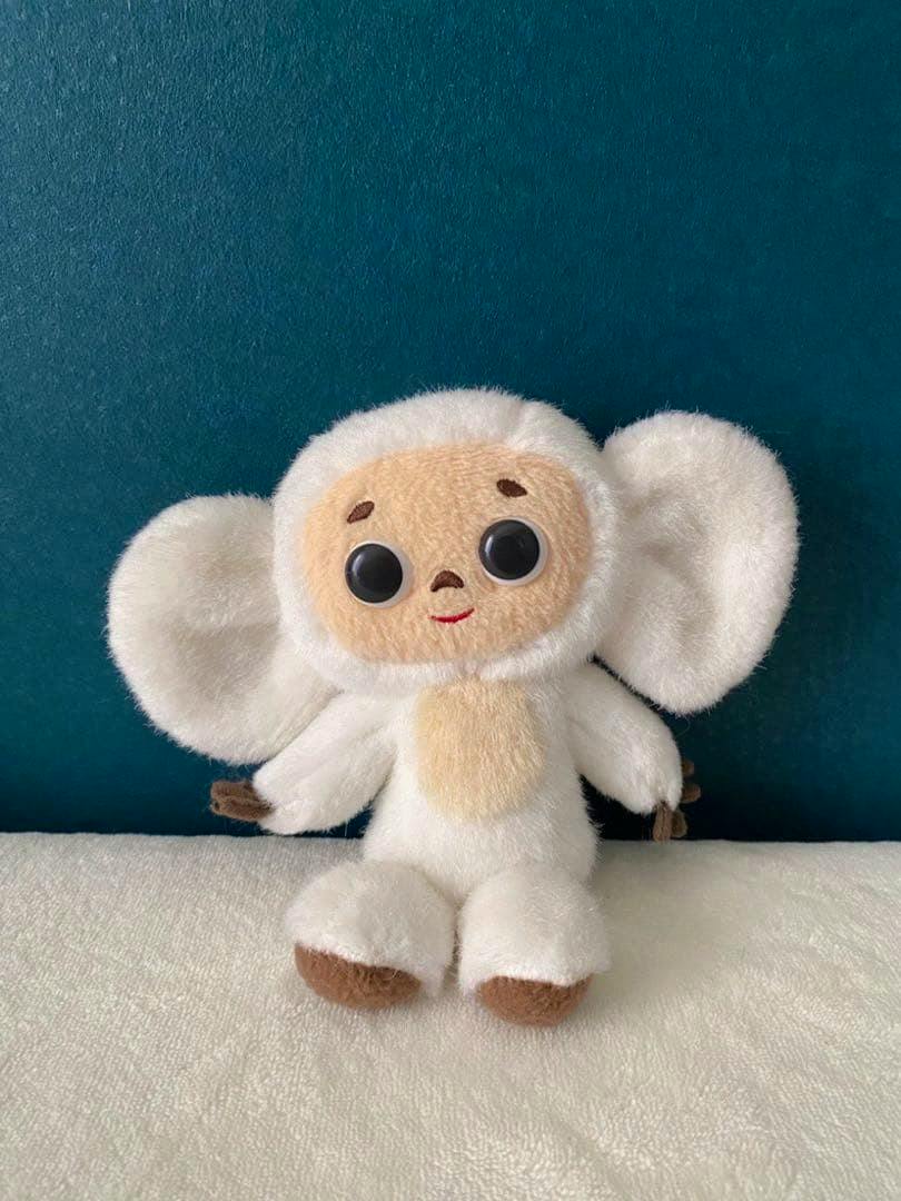 Front view of a Cheburashka plush toy with large round ears, a beige face, and black button-like eyes sitting against a teal background.