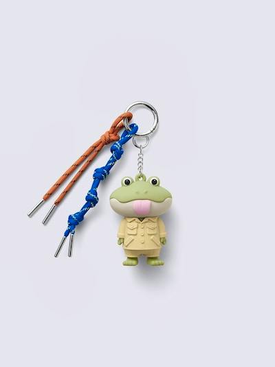 A plastic charm showing a green creature in a beige coat, attached to a silver keyring with blue and orange cords on a pale background.