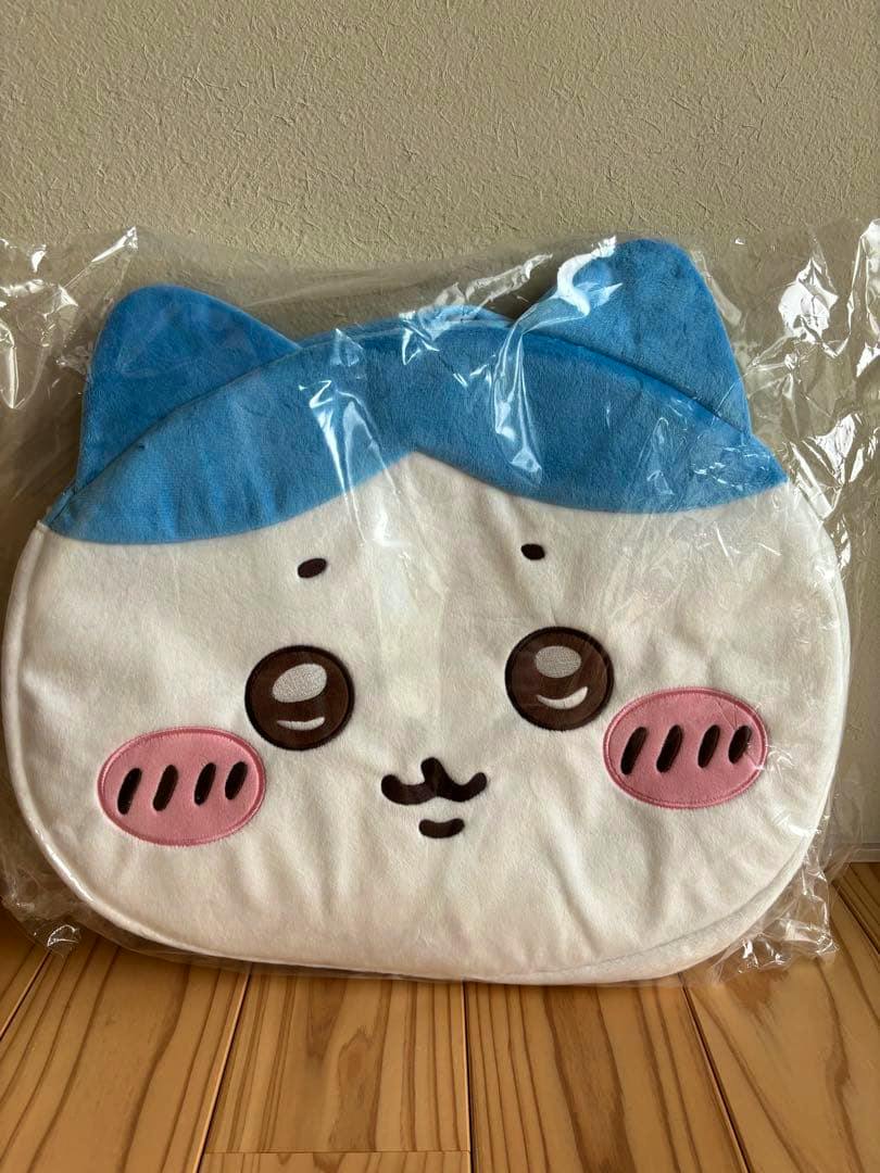 Front of the ちいかわ ハチワレ face bag showing blue ears and top, embroidered eyes, pink cheeks and mouth, photographed in its plastic packaging on a wooden floor.