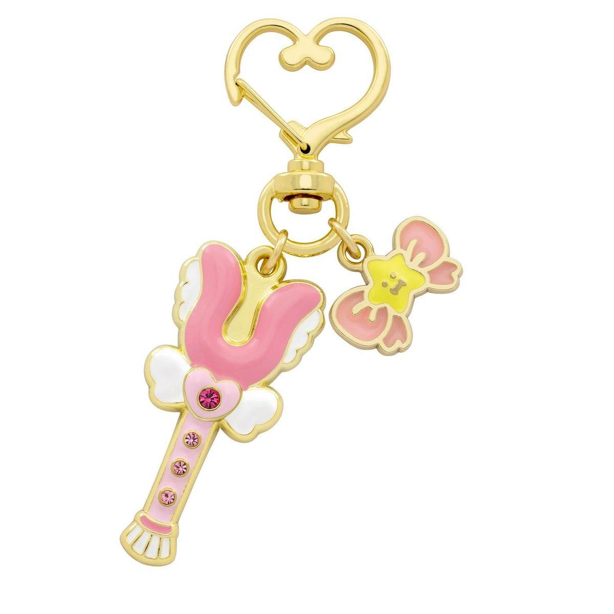 Gold metal keychain charm showing a pink magical stick with white wings and a small pink-and-yellow bow charm attached to a heart-shaped clasp.