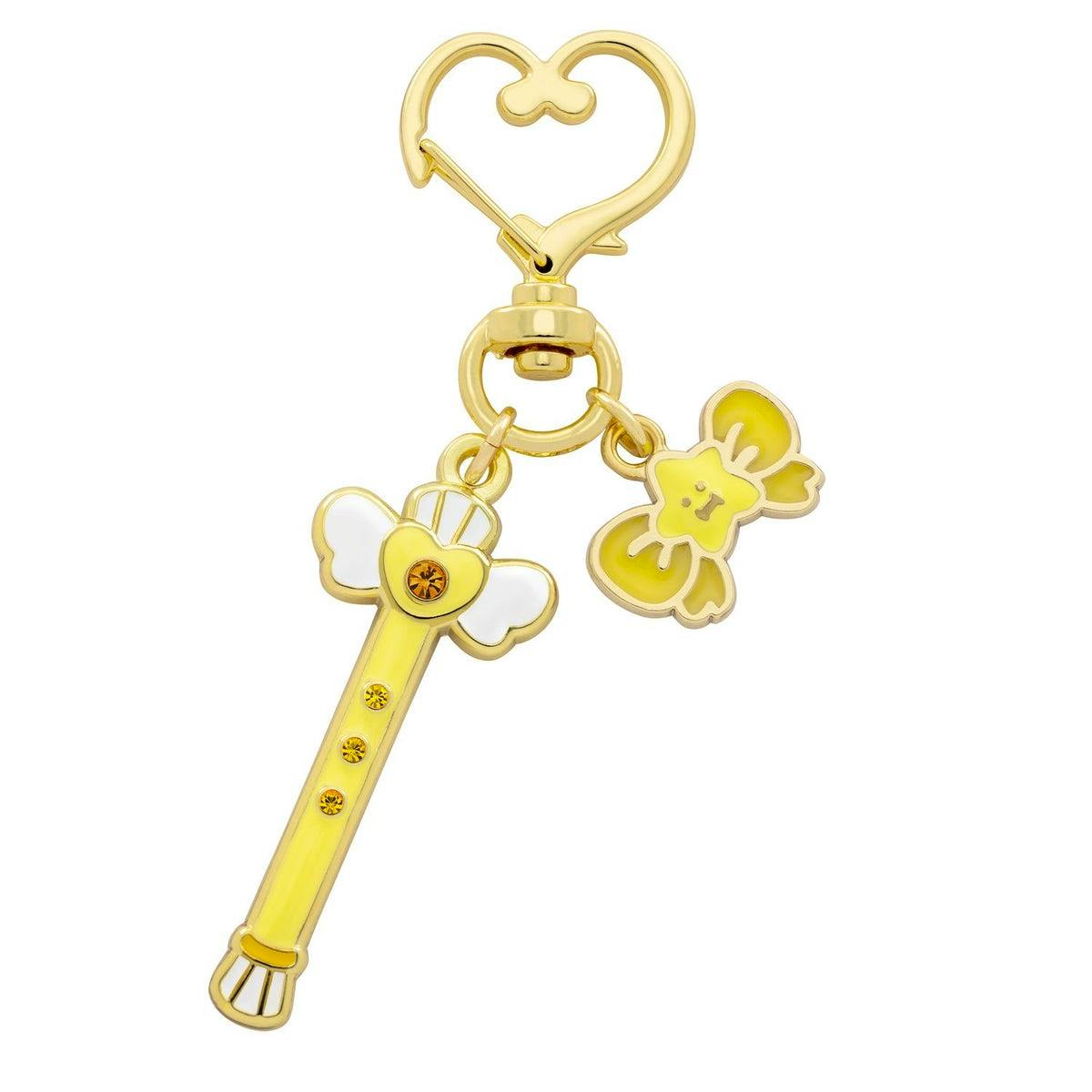 Close-up of a yellow-gold metal magical stick keychain charm with a heart-shaped clasp and a small bow charm attached.