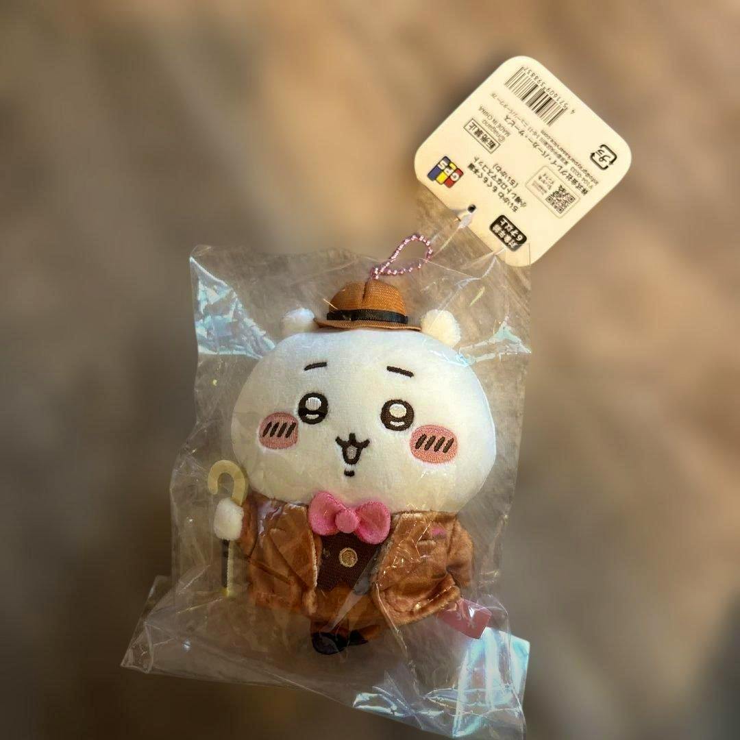 Plush ちいかわ mascot wearing a brown retro jacket, pink bow tie, and small hat, sealed in clear plastic with a white tag.