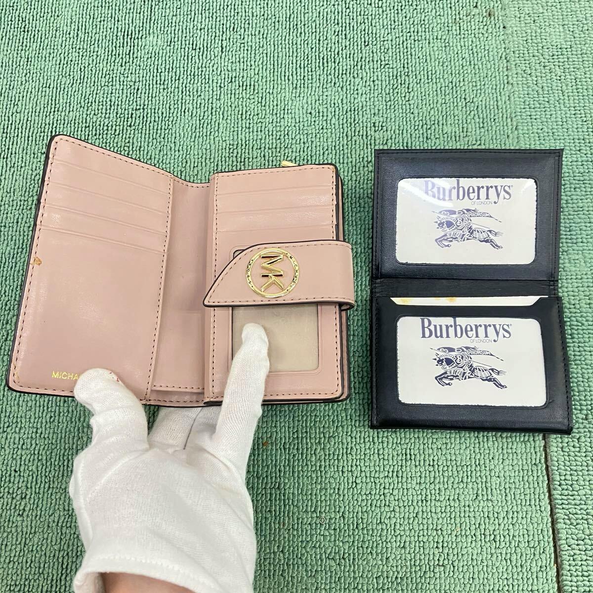 Open pink MICHAEL KORS wallet (wallet, cow leather) showing card slots and an ID window on a green mat with a gloved hand pointing at it.