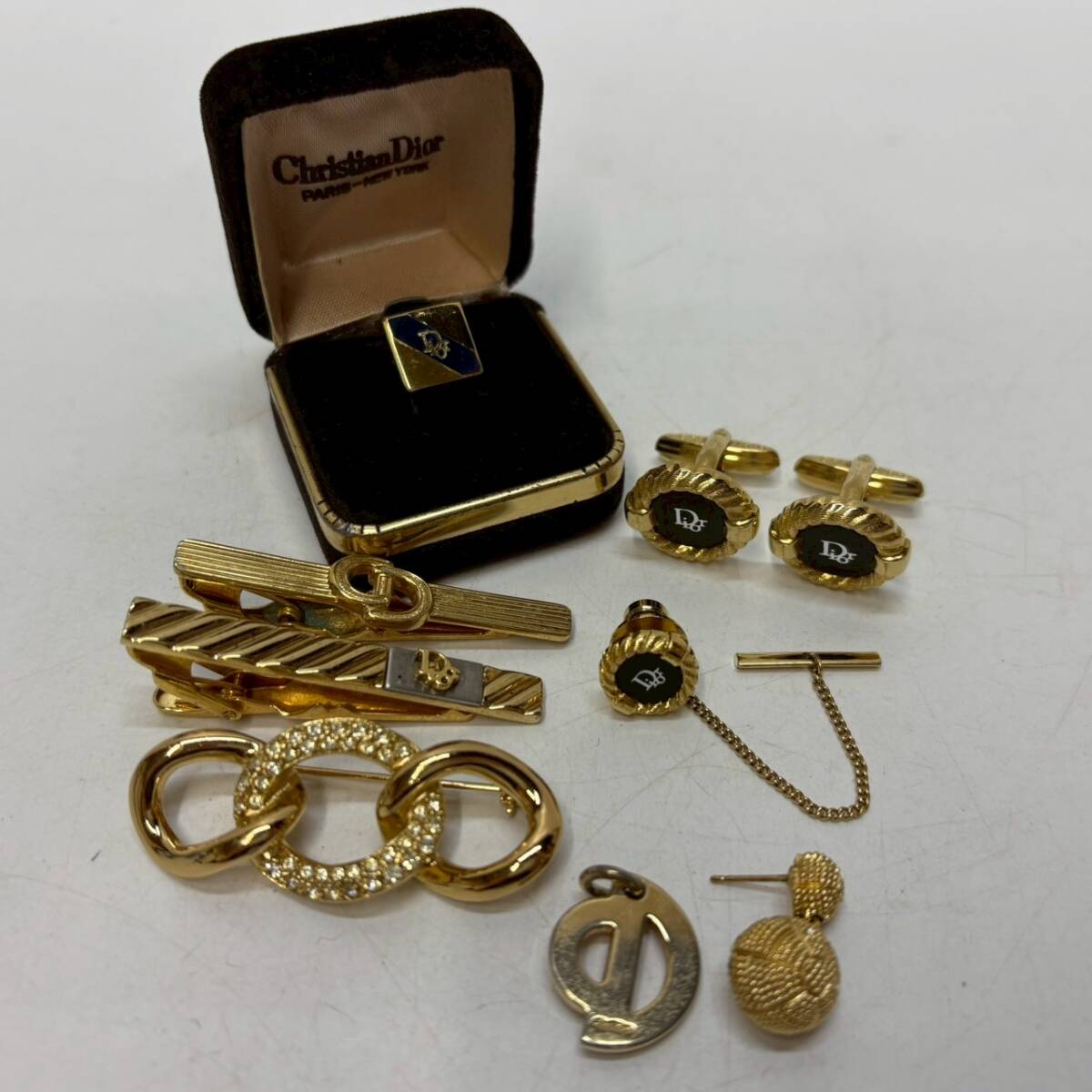 Assorted gold-tone Christian Dior accessories including cufflinks, tie bars, a brooch, and a boxed pin displayed on a white surface.