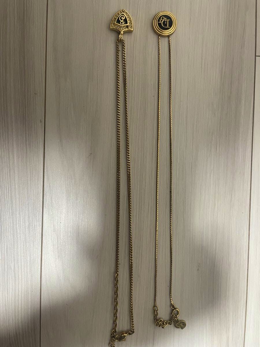 Two gold-color necklaces laid flat on a light wood floor, showing logo pendants and chains.