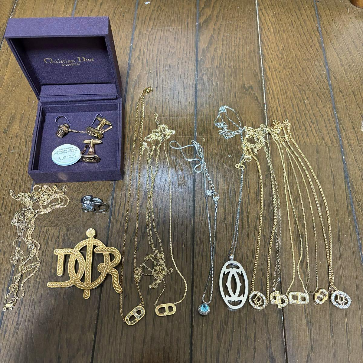 Top-down photo of assorted necklaces, pendants, a brooch, and a Christian Dior box arranged on a wooden floor.