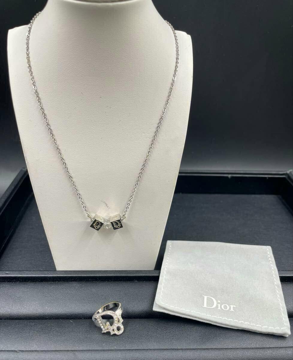 Silver-tone Christian Dior necklace with two cube (dice) CD logo charms displayed on a white bust, with the matching ring and a Dior pouch beside it.