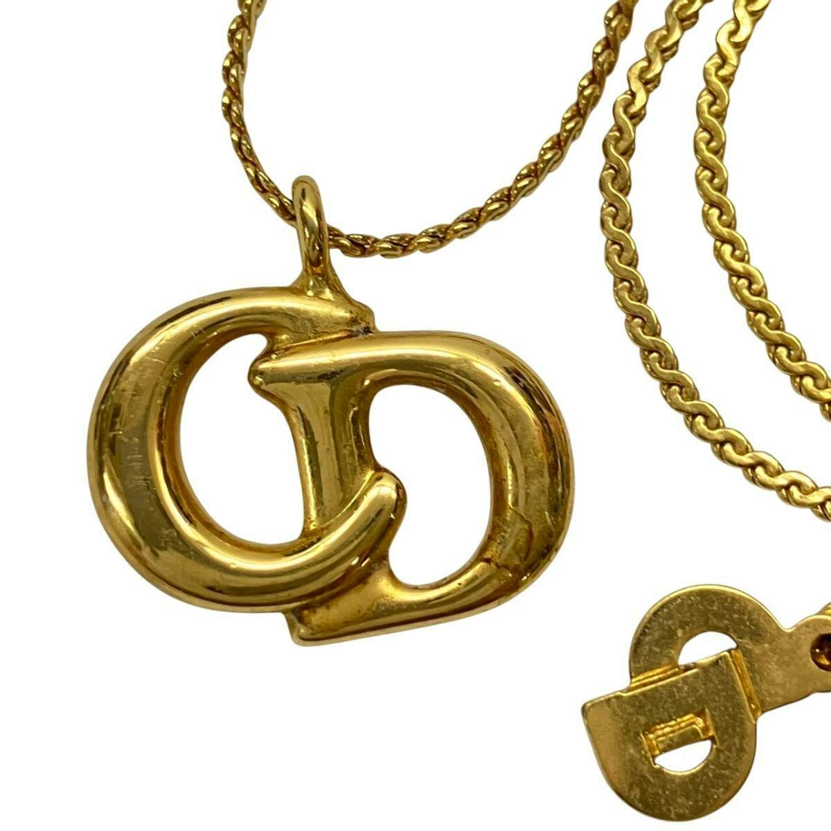 Close-up of a gold-colored Christian Dior CD logo pendant with part of its chain visible.