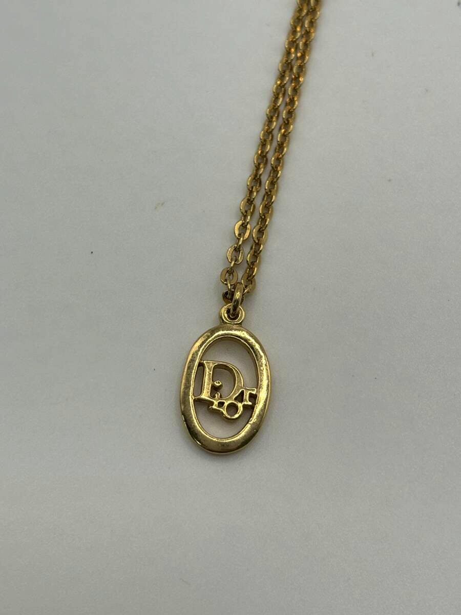 Gold-colored oval Dior pendant with a CD logo hanging on a short chain on a plain light background.