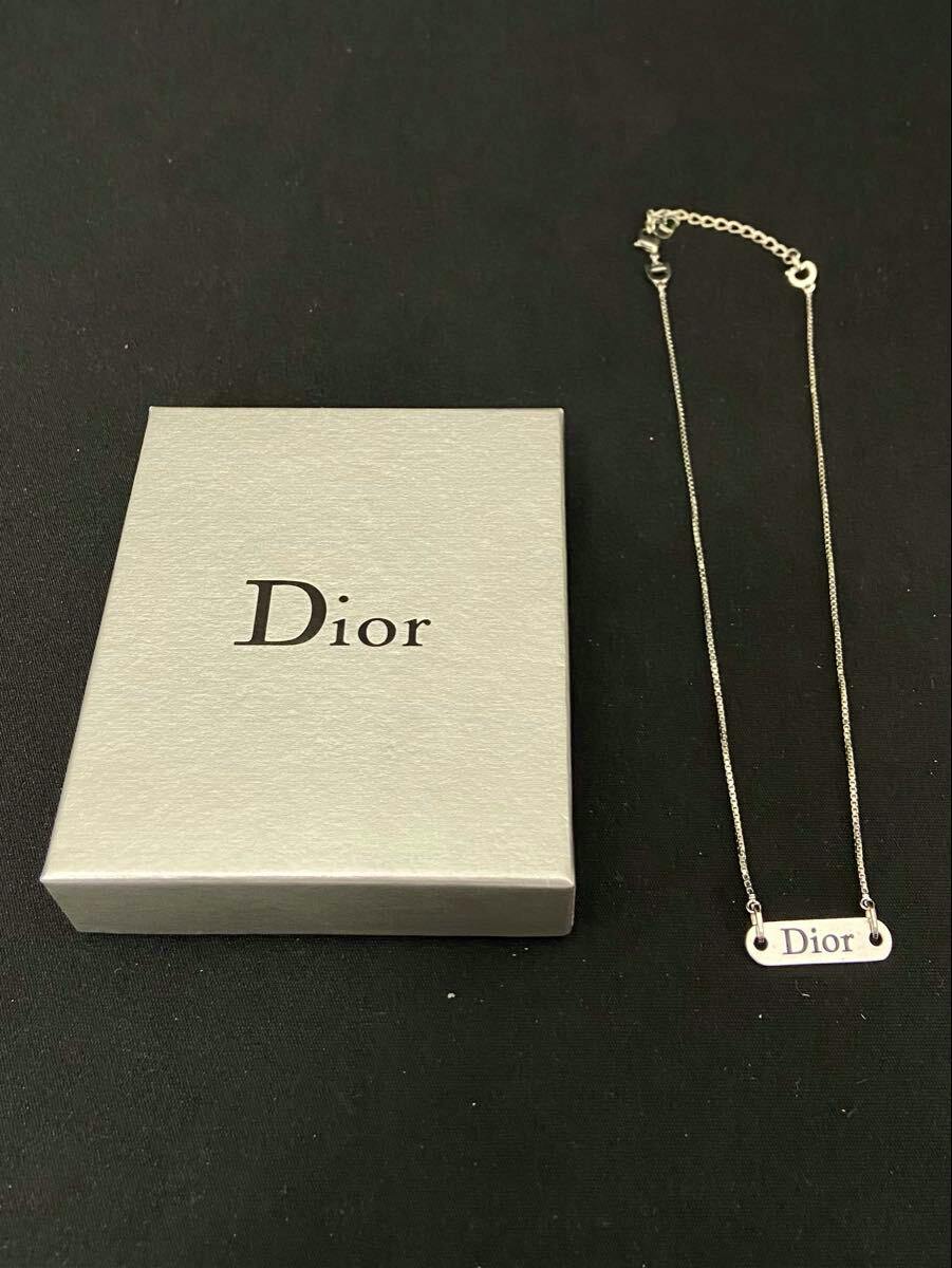 Silver-color Christian Dior plate-logo necklace next to a Dior box on a black background.