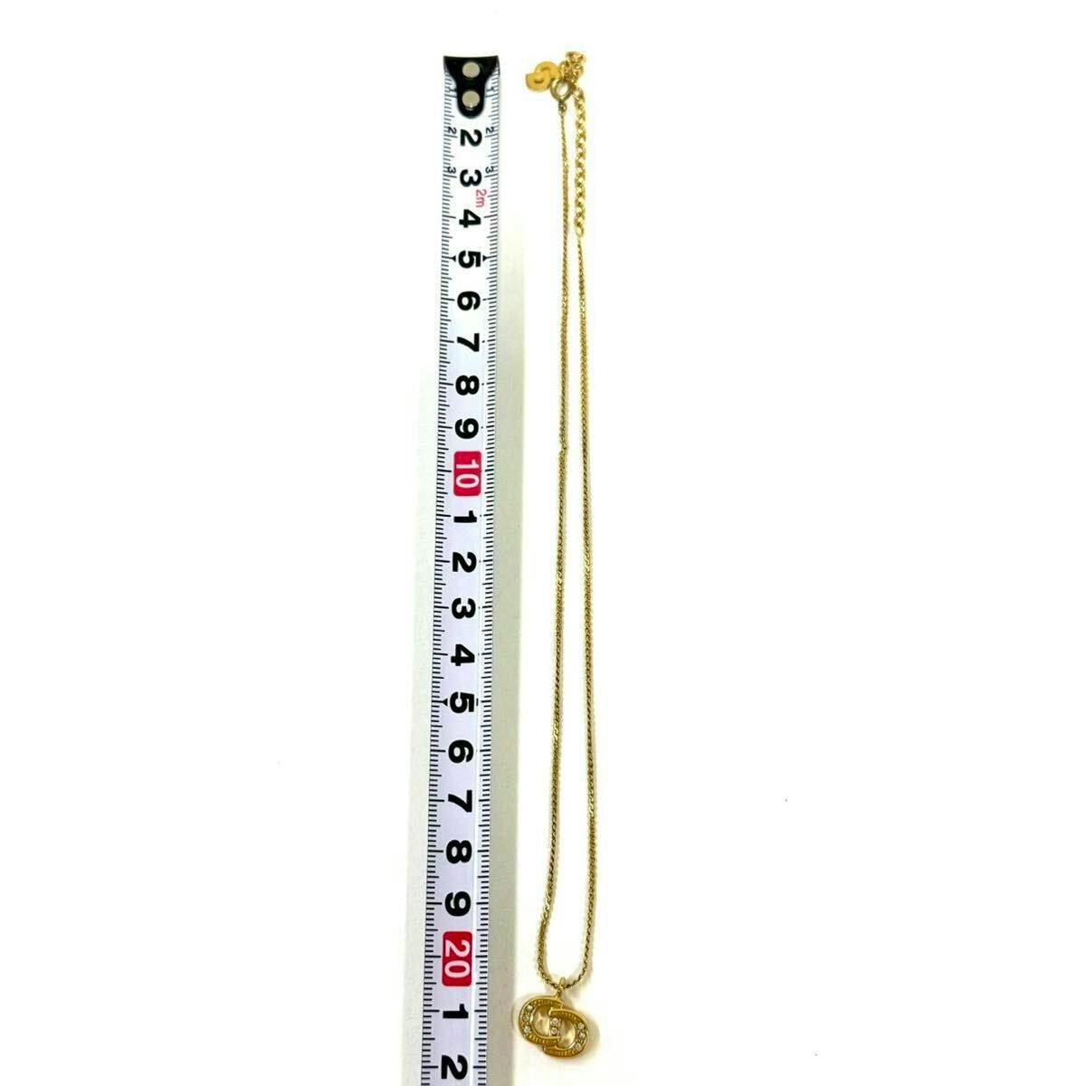 Gold-colored Christian Dior CD logo necklace with rhinestone pendant displayed beside a measuring tape on a white background.
