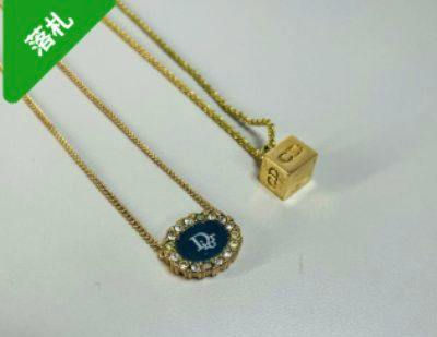 Two gold-colored Christian Dior necklaces on a white background: one oval rhinestone-bordered pendant with a CD logo and one cube pendant with engraved letters.
