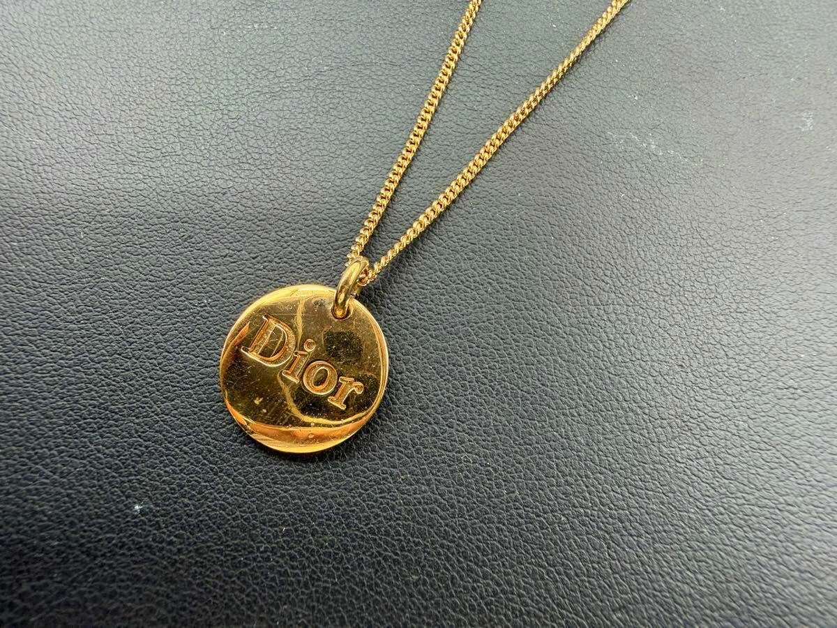 Gold-colored Christian Dior necklace with a round circle plate pendant engraved “Dior” on a dark textured surface.