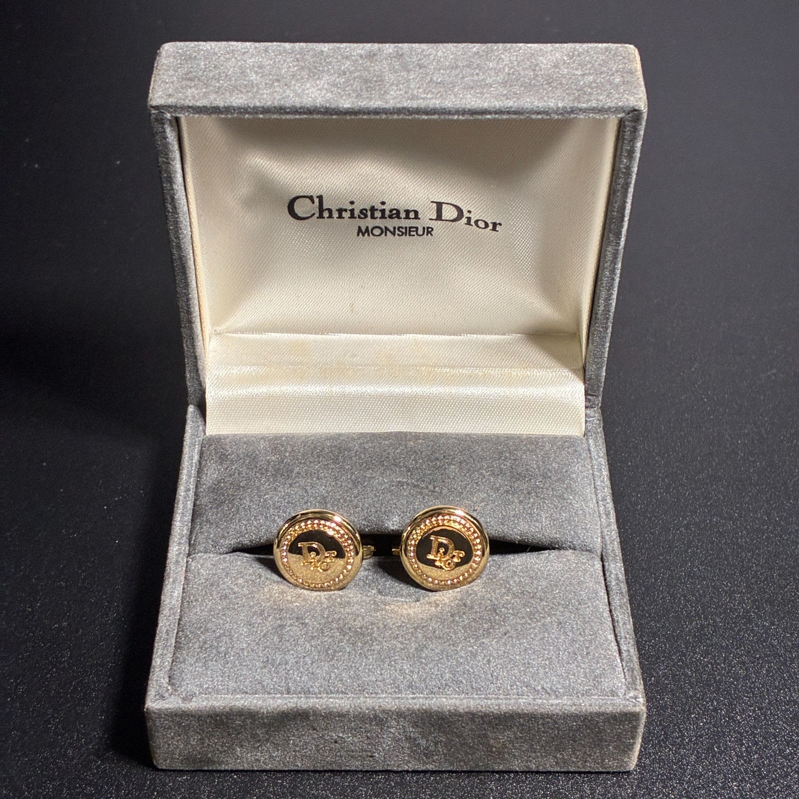 A pair of gold-tone Christian Dior cufflinks displayed inside a gray velvet presentation box labeled Christian Dior MONSIEUR.