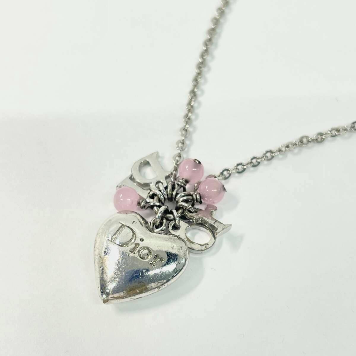 Silver-color Christian Dior necklace on a white background with a heart-shaped pendant engraved “Dior”, Dior letter charms, and pink bead accents on a chain.