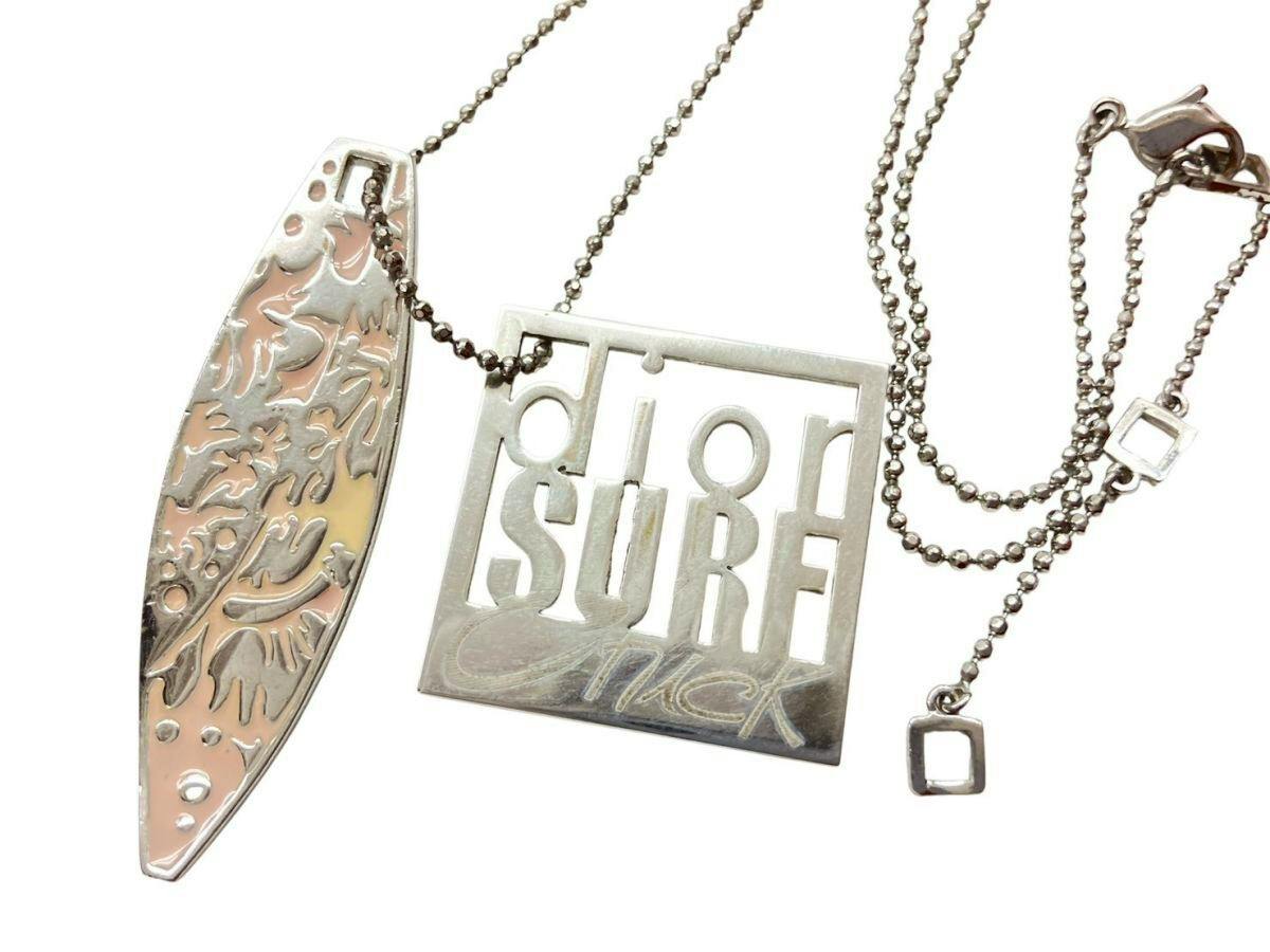 Close-up photo of the Christian Dior silver-colored necklace showing a square "SURF" logo plate, a surfboard-shaped pendant, and a ball chain with clasp.