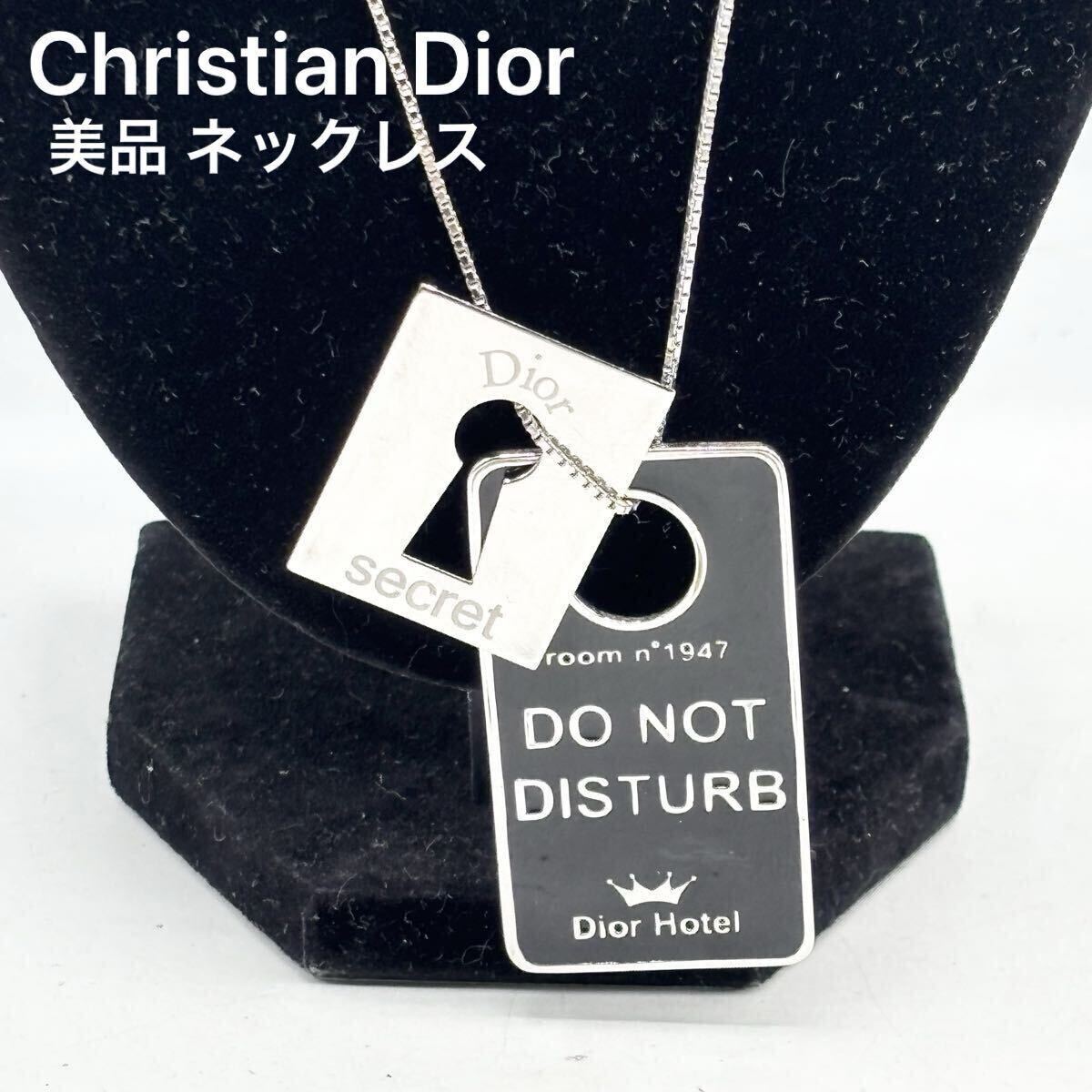 Christian Dior silver necklace with a keyhole-shaped plate and a black "DO NOT DISTURB" plate charm displayed on a black necklace stand.