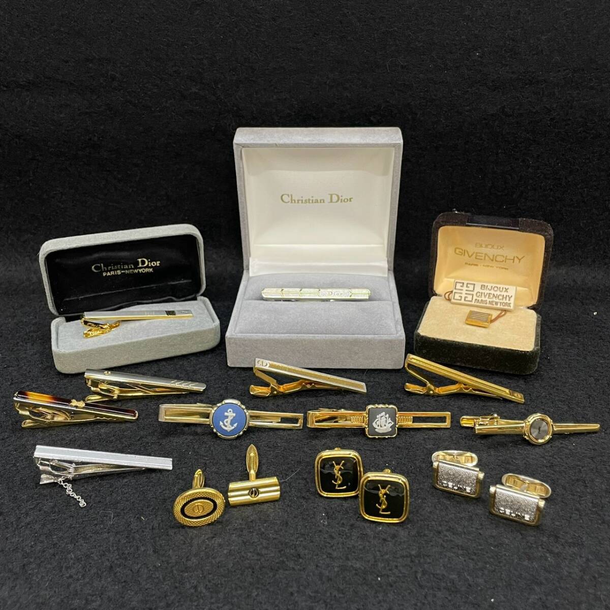 A collection of tie pins, tie clips, and cufflinks arranged on a black surface with Christian Dior and Givenchy boxes.