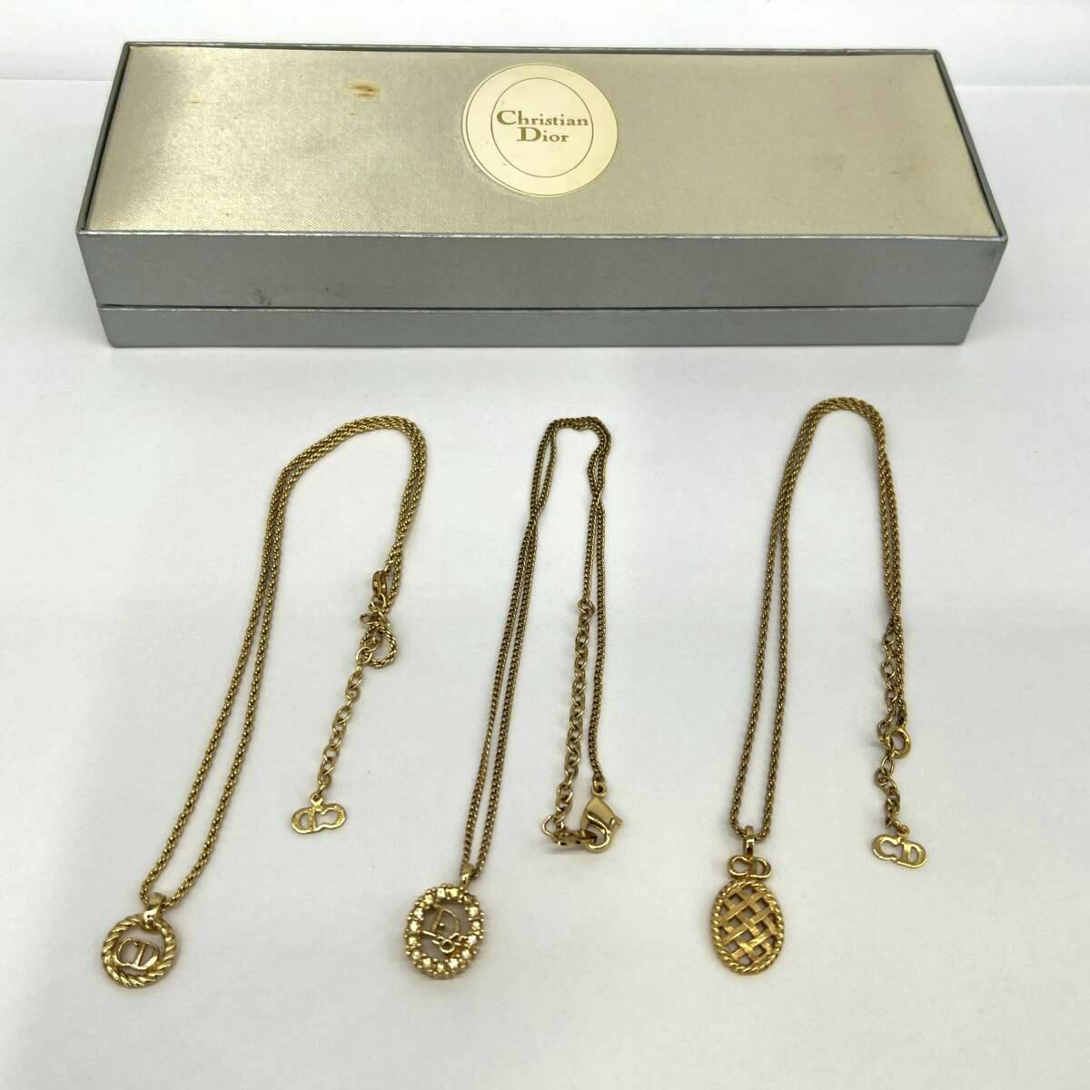 Three gold-colored Dior necklaces displayed on a white surface with a Christian Dior box behind them.