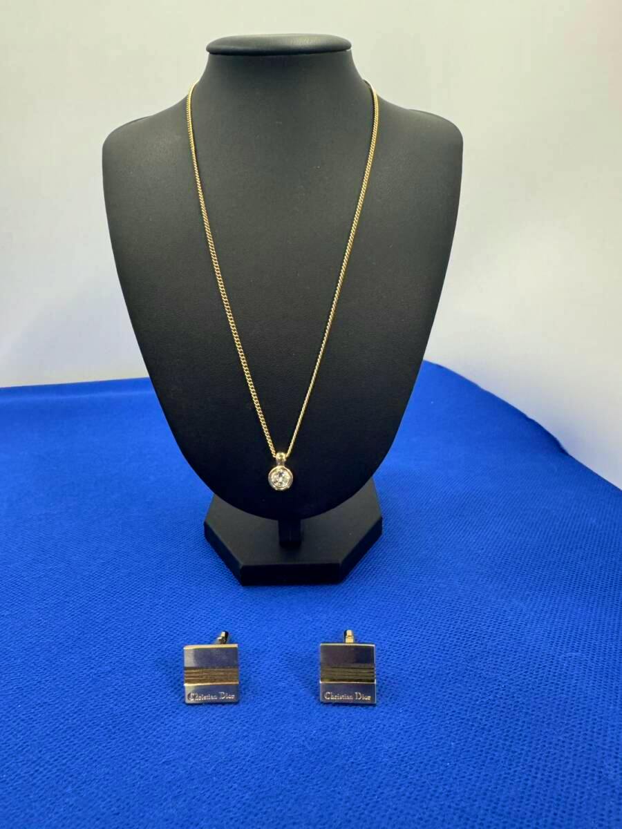 Gold-color Christian Dior necklace with a clear stone pendant on a black display bust, with matching square gold-color cufflinks on blue fabric.