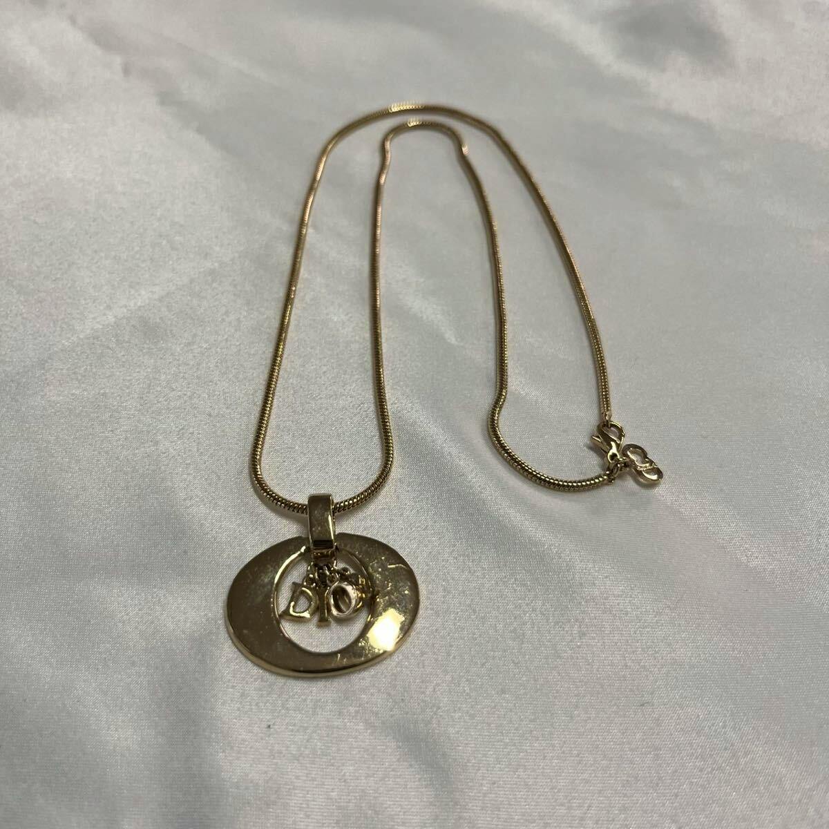 Gold-tone Christian Dior necklace with a circular pendant and small "Dior" letter charms laid flat on white fabric.