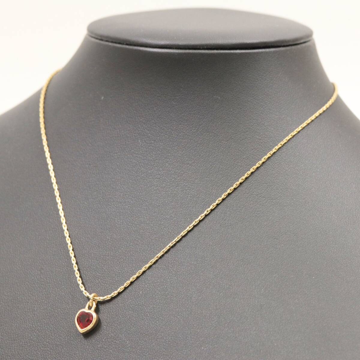 Gold-colored chain necklace with a small red heart pendant shown on a black display bust.