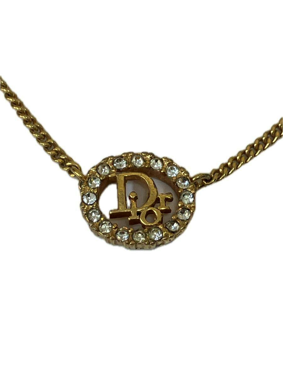 Close-up of a gold-tone necklace pendant showing an oval rhinestone-studded frame with a Dior logo and attached chain.