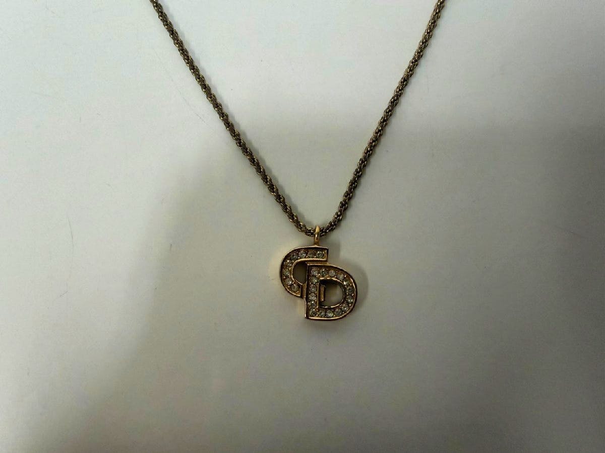 Photograph of a Christian Dior necklace showing a rhinestone-studded "CD" logo pendant on a twisted rope chain against a white background.