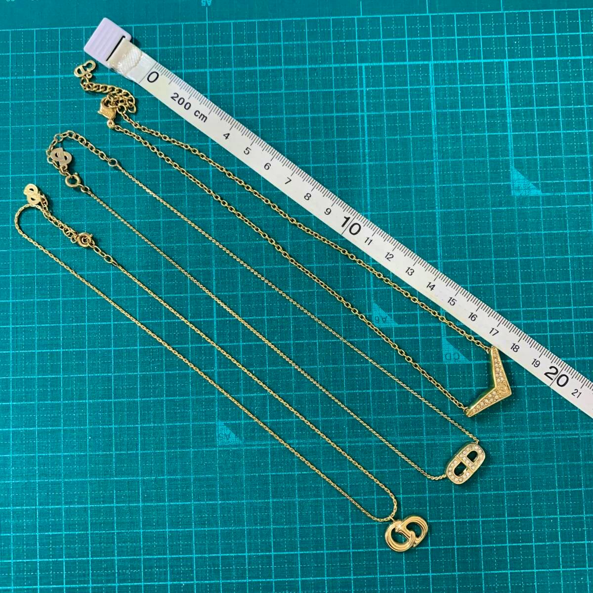 Three gold-tone Christian Dior necklaces with different pendants laid flat on a teal grid cutting mat next to a measuring tape.