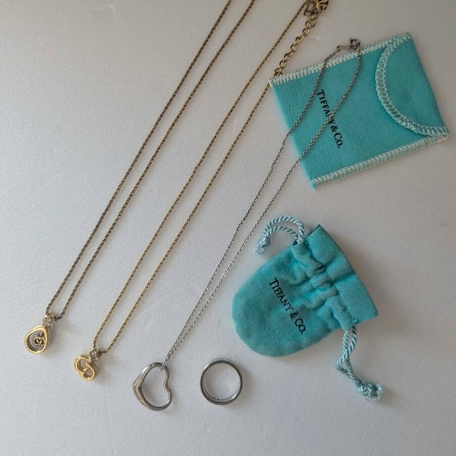 Two gold chains with small pendants, a silver heart pendant necklace, and a silver ring displayed with Tiffany & Co. pouches.