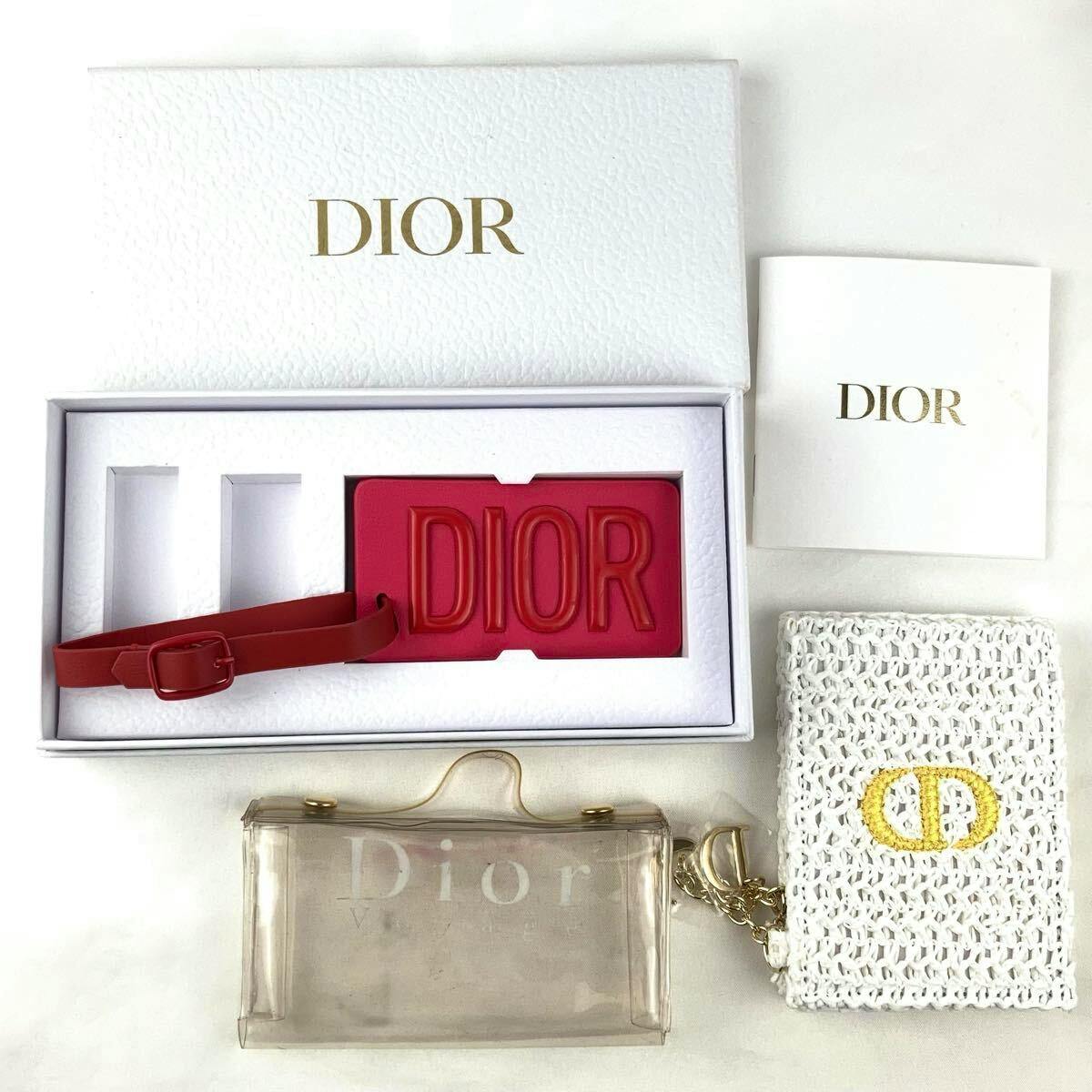 Top-down flat lay showing a red DIOR name tag in a white DIOR box, a clear vinyl Dior pouch, a white woven pouch with a gold CD logo, and a small DIOR card.