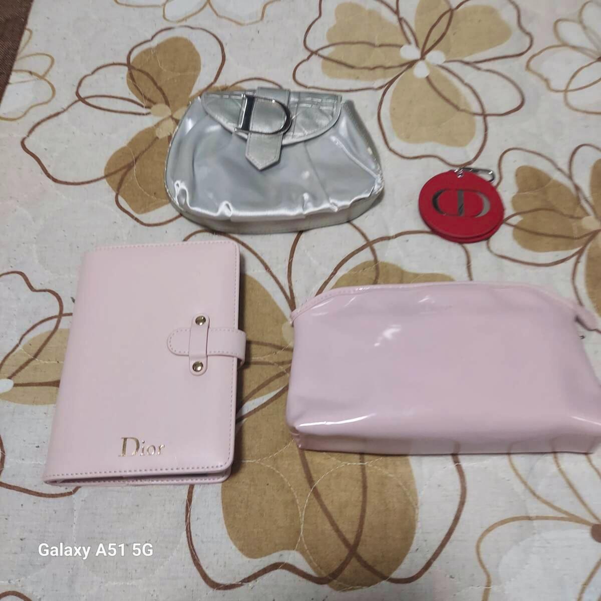 Bundle of Dior items on fabric: a pink Dior notebook/wallet, a pink patent pouch, a silver pouch, and a red round compact mirror.