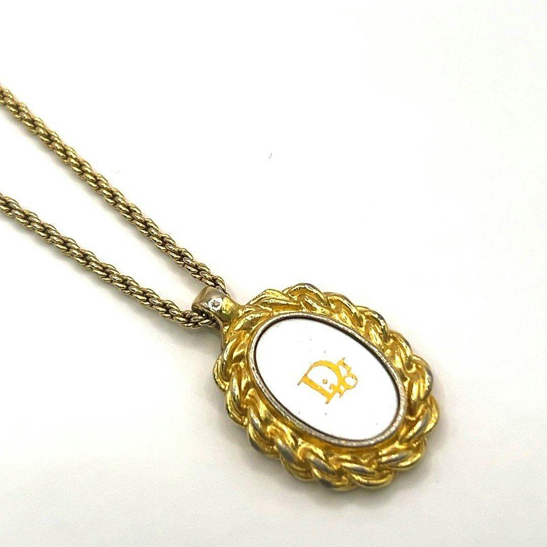 A Christian Dior necklace laid on a white background showing an oval pendant with a white center and Dior logo on a gold-tone chain.