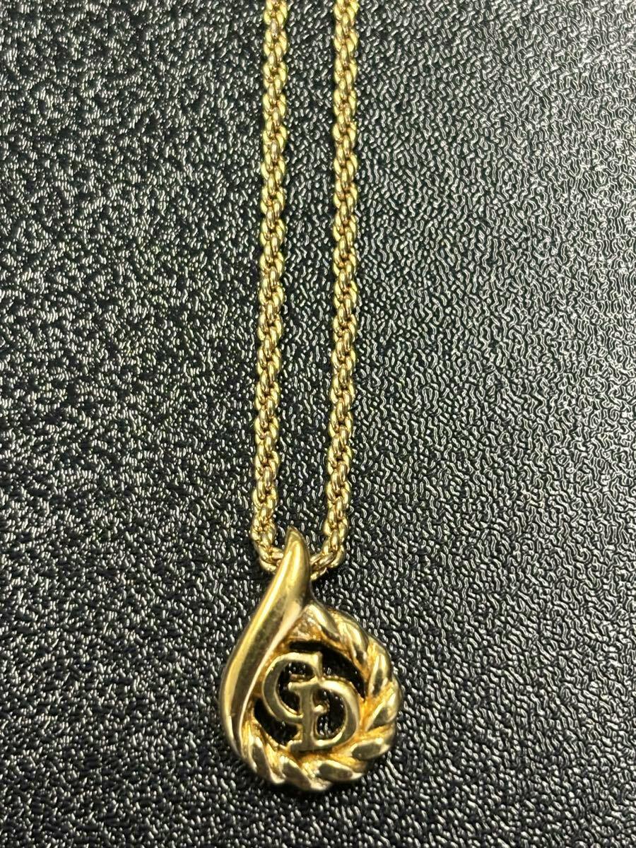 Christian Dior gold leaf pendant with interlocking CD logo on a gold chain lying on a black textured surface.