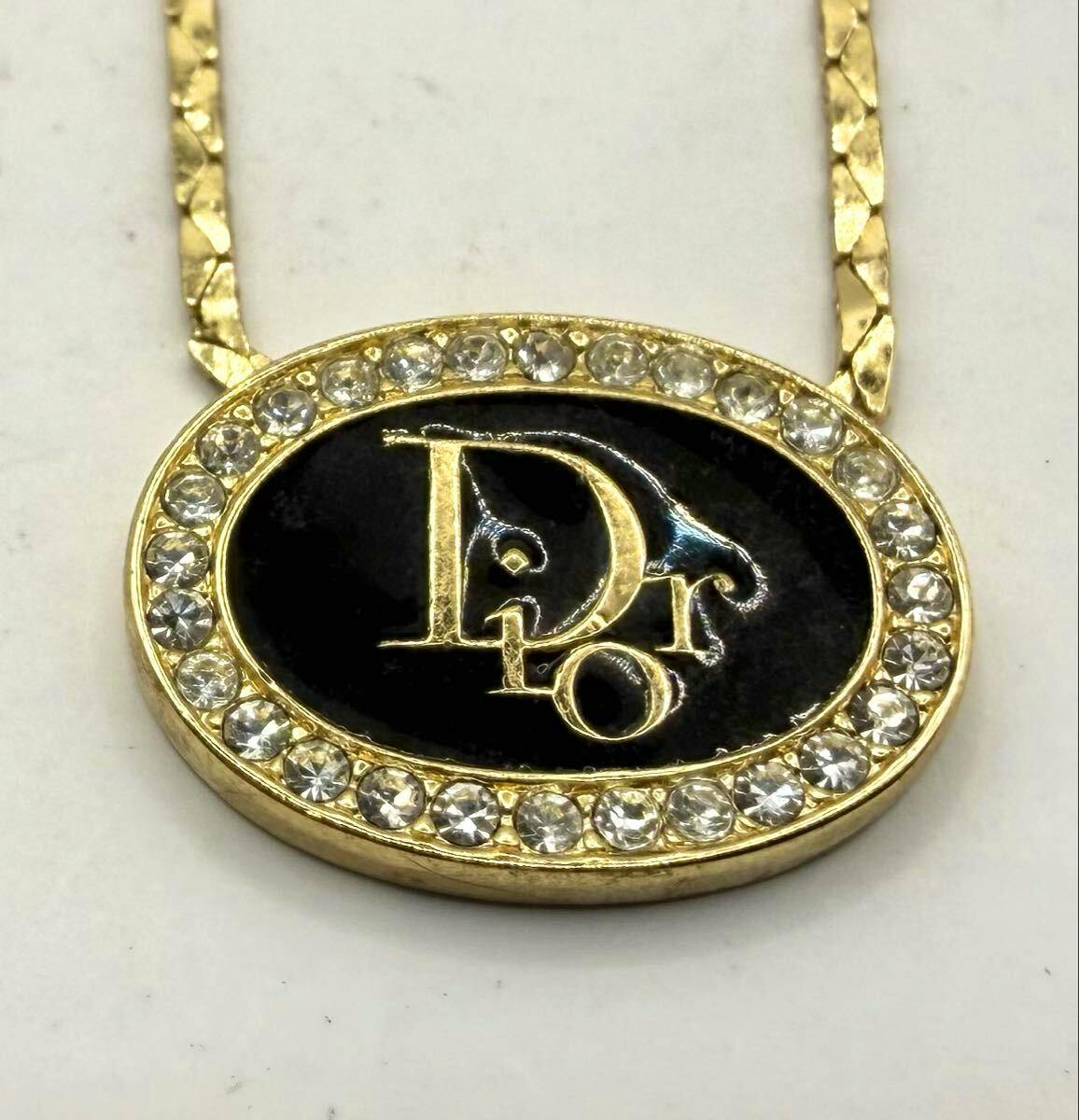 Close-up of a Christian Dior gold-tone oval pendant on a chain, with a black enamel center, gold Dior logo, and a rhinestone border.
