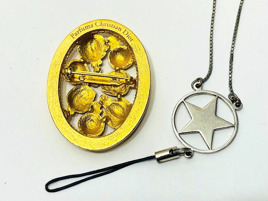 Close-up of a gold oval Christian Dior brooch and a silver star pendant necklace with strap on a white background.