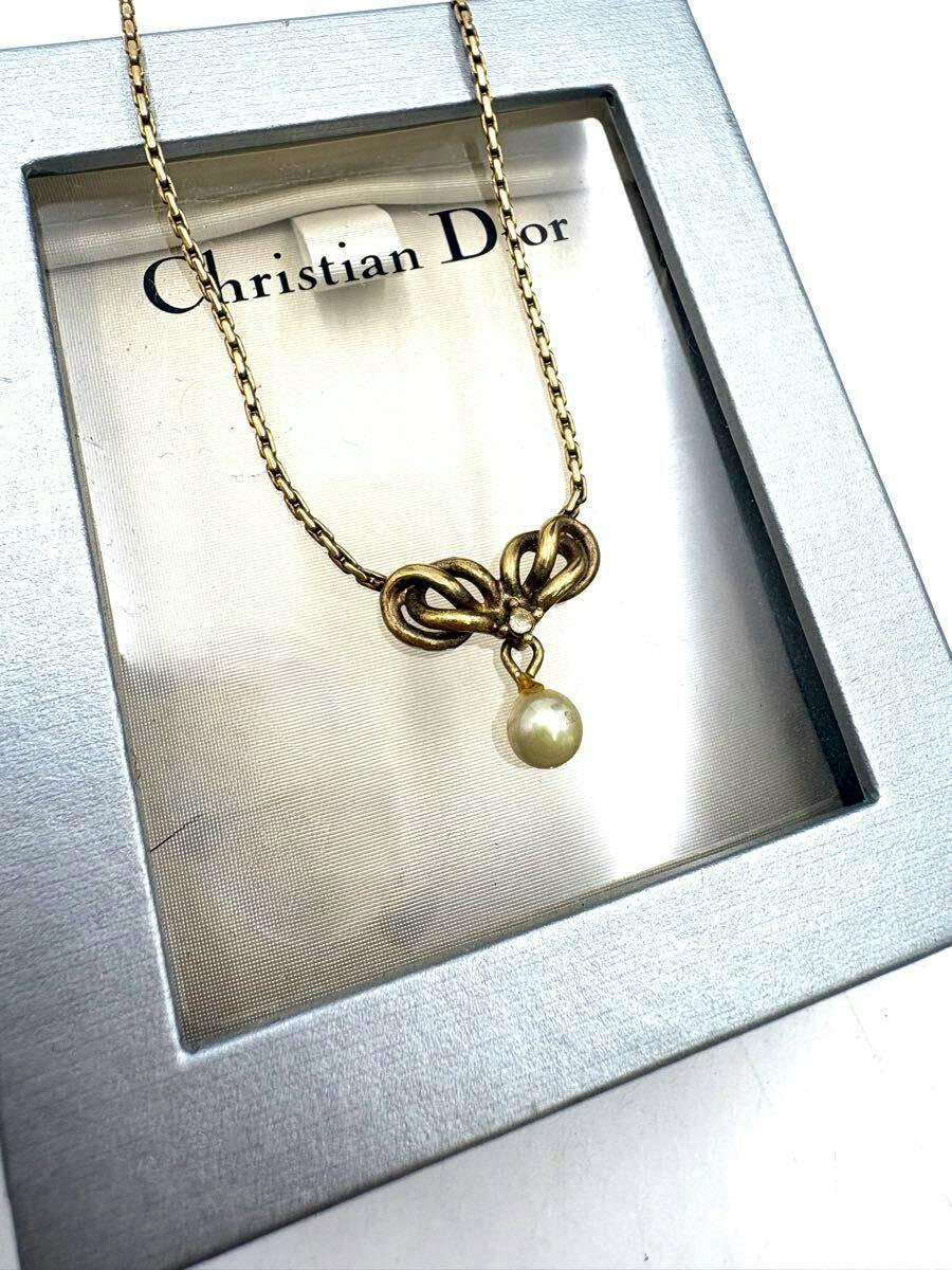 Gold-colored Christian Dior necklace with a ribbon-shaped pendant and a faux pearl displayed in its box.