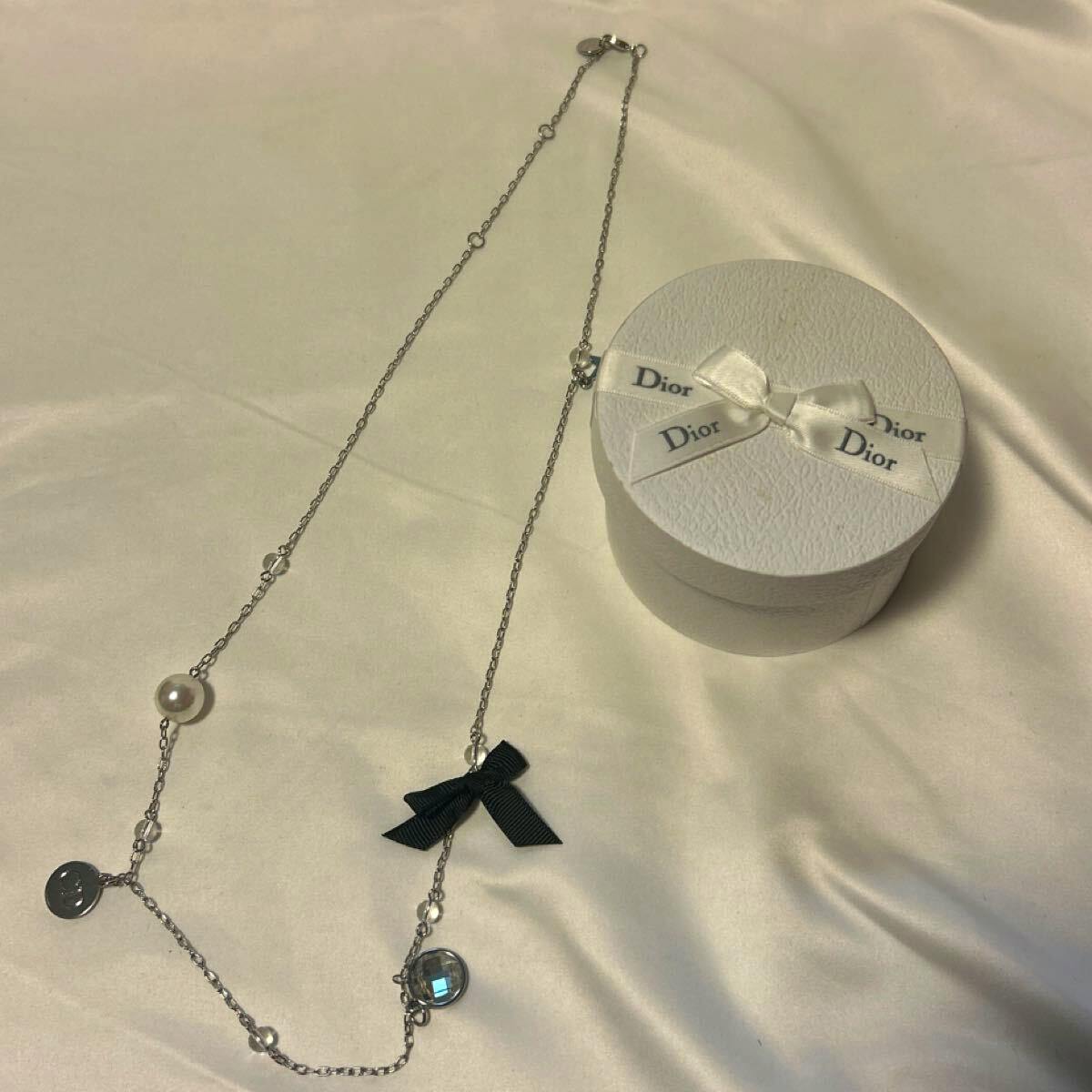 Long silver-colored necklace with a pearl, charms, and a black ribbon shown next to a round Dior box.