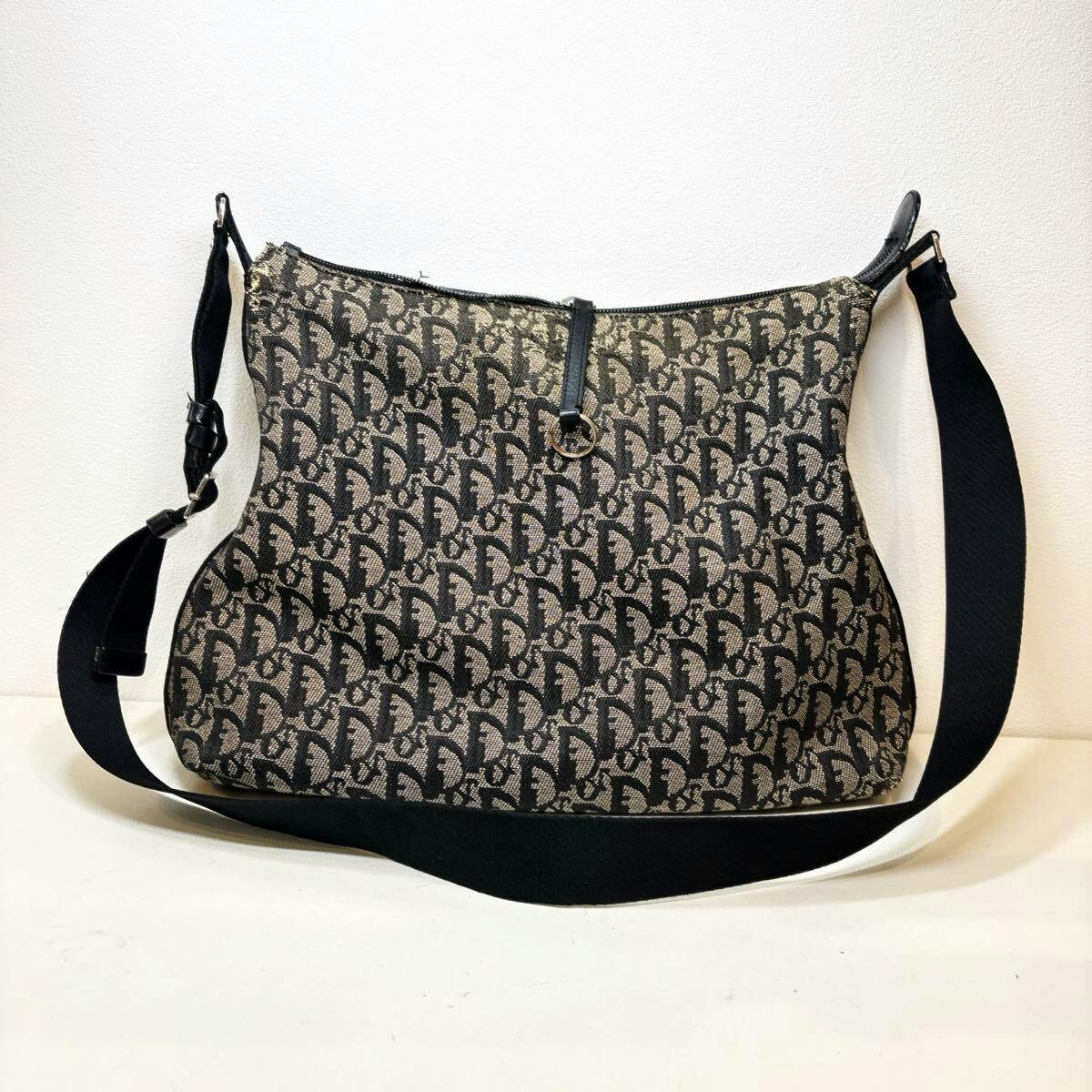 A Christian Dior canvas Trotter shoulder bag in black with all-over monogram and an adjustable black strap against a white background.