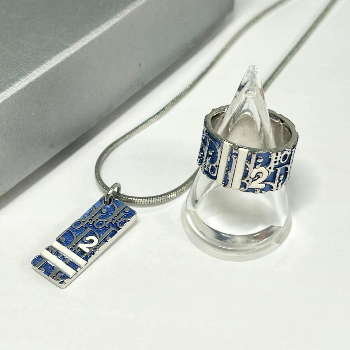 A blue Christian Dior trottoir-pattern ring displayed on a clear cone next to a matching rectangular trottoir-pattern pendant on a silver chain.