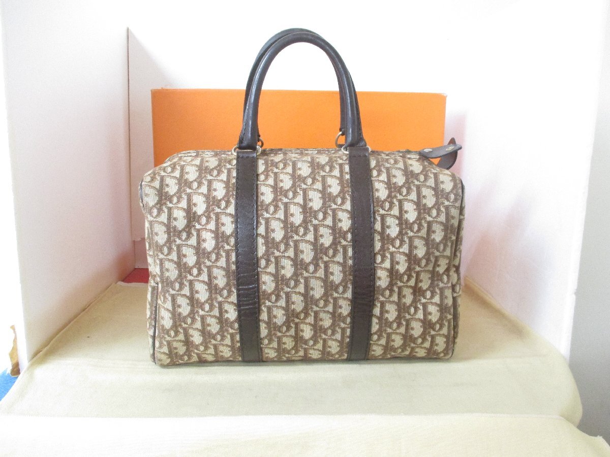 Front view of a brown Dior Trotter monogram vintage Boston handbag with dark leather handles, placed in front of an orange box.