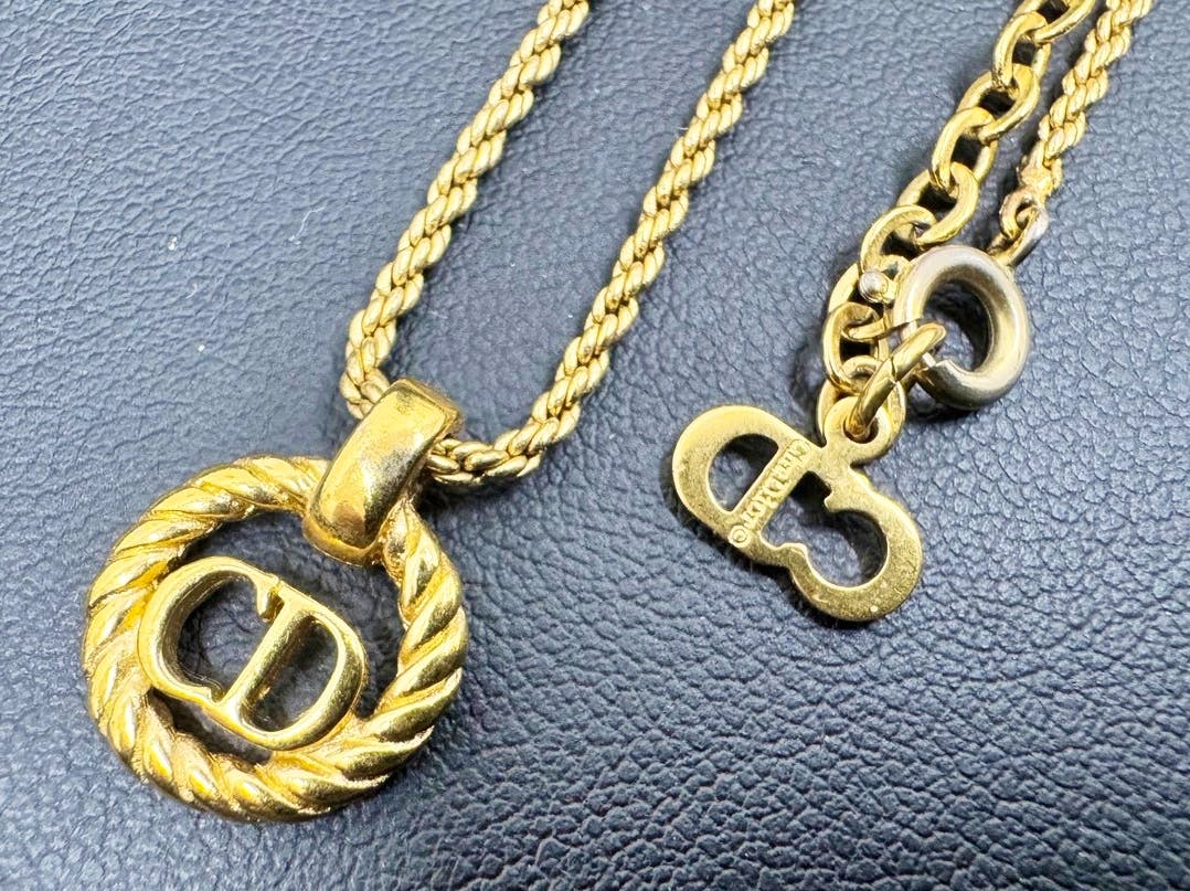 Close-up of a Christian Dior gold-tone rope chain necklace with a twisted circle CD logo pendant and a nearby chain tag on a black textured surface.