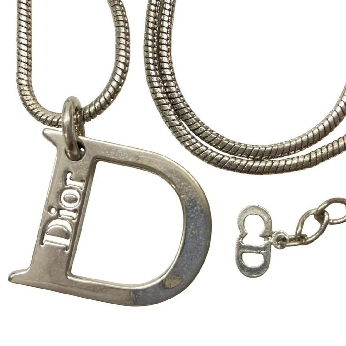 Close-up of a silver-colored Christian Dior D-logo pendant and part of its chain on a white background.